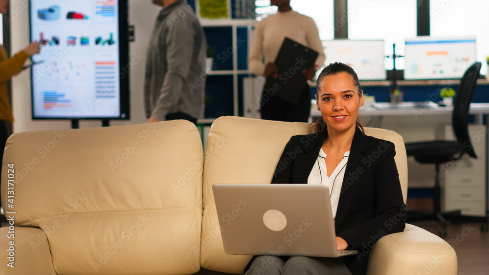 Poster hispanic business woman smiling at camera sitting on couch typing on computer while diverse colleagu