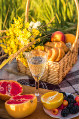 Picnic basket with food and glass with champagne on green sunny lawn. there are two croissants, grapefruit, orange, berries, peaches.