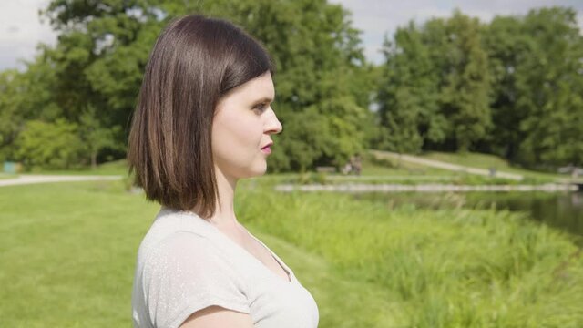 A young Caucasian woman looks around thoughtfully by a pond in a city park on a sunny day - trees and a pathway in the blurry background