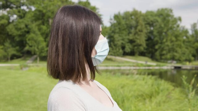 A young Caucasian woman in a face mask looks around thoughtfully by a pond in a city park on a sunny day - trees and a pathway in the blurry background