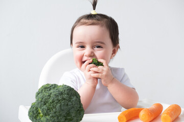 Baby girl sitting in baby chair eating carrot and broccoli on white background.