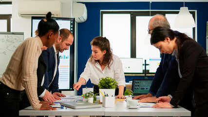 Diverse team of workers talking, looking at documents and analyzing graphs data standing at conference desk in start up company. Professional colleagues team discussing marketing project brainstorming