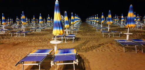 Panorama of empty beach at night with blue and yellow umbrellas and sun loungers.