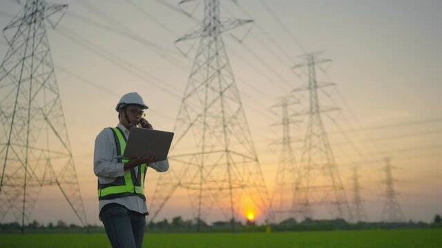 An electrical engineer with a laptop talking on the phone High voltage tower inspection report before starting a project Assigned by the organization during the time of sunset or sunrise.