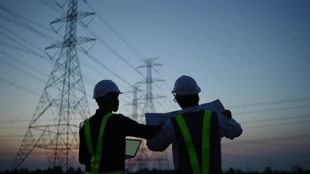 Two electrical engineers discussing a project on a high-voltage tower in the evening.
