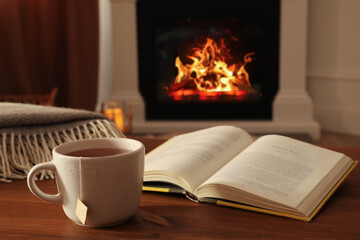 Cup of hot tea and book on wooden table near fireplace at home. Cozy atmosphere