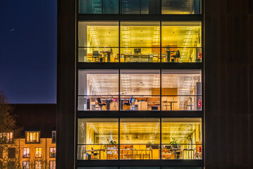 A contemporary office with a broad modern, glazed facade illuminated at night. Square pattern of wide glazed windows of a financial firm exterior. Tech industry headquarter with office space interiors