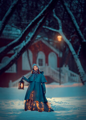 Girl in the blue cape standing at the night snowy winter park with lantern