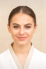 Portrait of a beautiful young caucasian woman with dark hair wearing white bathrobe looking at camera and smiling on a beige background