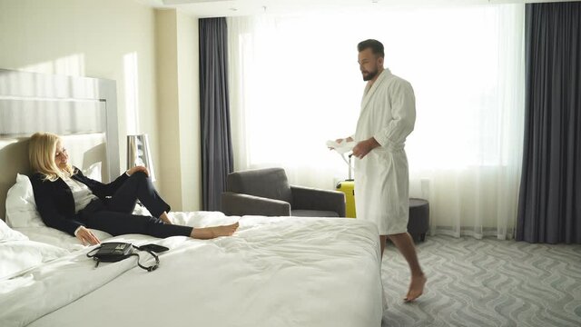 Female in suit sits on bed waiting for boyfriend after shower, shirtless guy stand looking at her, flirting.