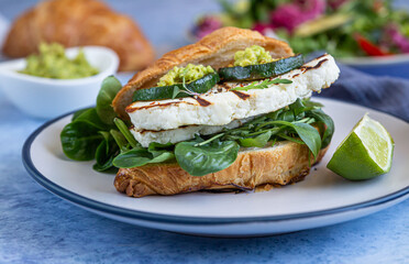 Croissant sandwich with grilled halloumi cheese, arugula and grilled zucchini, blue concrete background. Healthy breakfast or lunch. Close up.
