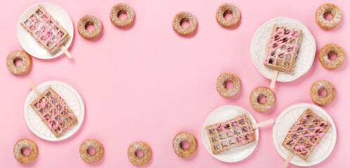 Traditional belgian waffles with sprinkles and delicious doughnuts with powdered sugar on pastel pink background.