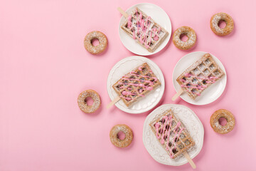 Traditional belgian waffles with sprinkles and delicious doughnuts with powdered sugar on pastel pink background.