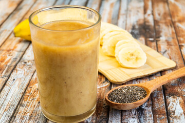 Glass of fresh Banana smoothie, served with open banana, spoon of chia seeds on wooden table. Selective focus