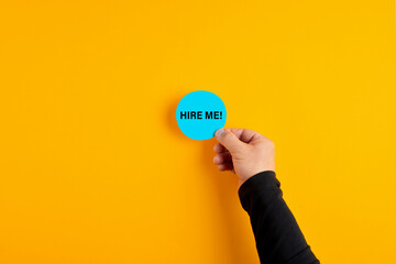 Male hand holds a blue badge with the word hire me on yellow background. - obrazy, fototapety, plakaty