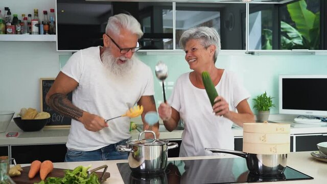 Happy senior couple having fun dancing together at home - Elderly people preparing health lunch in modern kitchen - Retired lifestyle family time and food nutrition concept