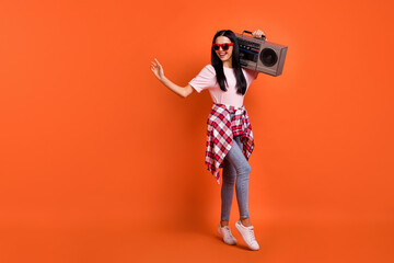 Full size photo of optimistic brunette lady stand wave hand hold boom box wear spectacles t-shirt jeans isolated on orange background