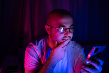 Young thoughtful man looking at his cellphone in blue and red neon light room