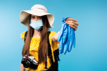 Fototapeta premium A blurred photo of a tourist girl in a hat and a medical mask stretching out latex protective gloves on the screen. Blue background. Pandemic and travel.