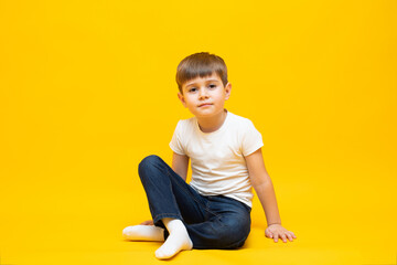 Naklejka premium Little boy in jeans and a white t-shirt sits on an isolated yellow background