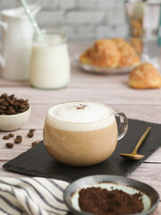 A cup of hot latte coffee with mousse, spoon & its bean in background with pot of milk and cookies. It's a beverage drink made with espresso and steamed milk