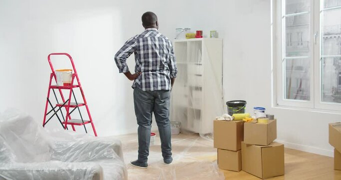 Rear of young adult African American handsome happy man standing in empty room with boxes looking at wall turning to camera in good mood, house renovation, renewing apartment, home repair concept