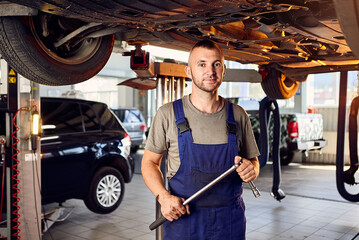 Handsome auto mechanic checking running gear of automobile on service station. Cheerful male worker...