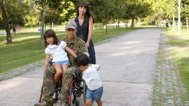 Disabled ex soldier walking with kids and wife in park. Woman wheeling wheelchair of husband, daughter sitting on dads lap. Veteran of war or returning home concept