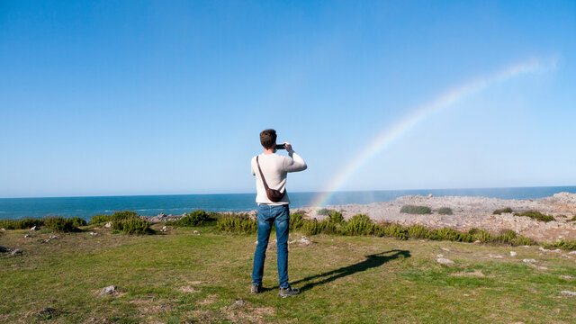 Hombre joven con vaqueros y jersey blanco fotografiando un arco iris en acantilado marino