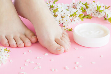 Obraz na płótnie Canvas Young adult woman feet. Opened white jar of natural herbal cream on light pastel pink background. Care about clean, soft and smooth skin. Beautiful branch of cherry blossoms. Fresh flowers. Closeup.
