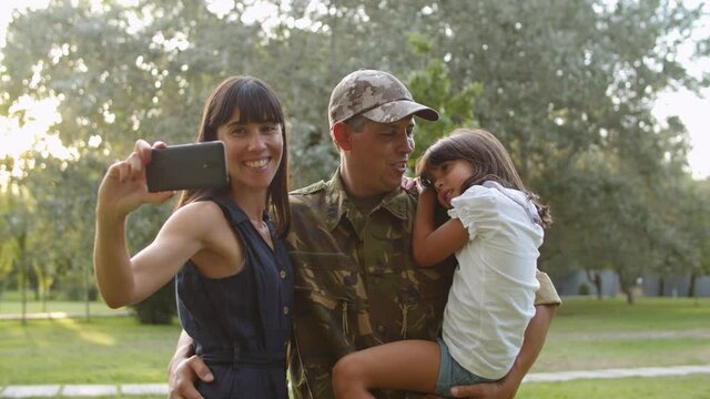 Happy military couple of parents and little daughter taking selfie on cellphone in city park. Soldier dad holding kid in arms and smiling at camera. Family reunion or returning home concept
