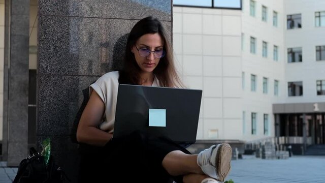 Freelance business woman in business attire works on a laptop in the park. Happy young girl relaxes, does internet surfing.