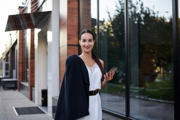 Young brunette girl with red pony tail, wearing stylish black jacket and white silk dress, standing near modern glass building, holding phone. Pretty business woman on lunch break. Female portrait