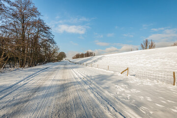 Fototapeta na wymiar Apparently endless long road at the foot of a Dutch dyke. It is winter and a thick layer of snow has fallen. Tire tracks are visible on the road and paw prints of wild animals are visible on the verge