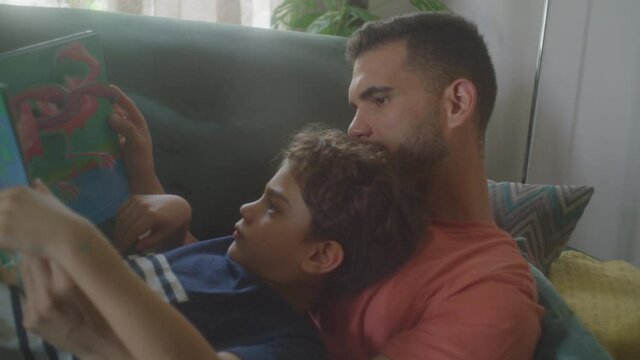 Father And Son Lying On The Couch At Home Watching A Movie, Playing And Having Fun On IPad Or Tablet