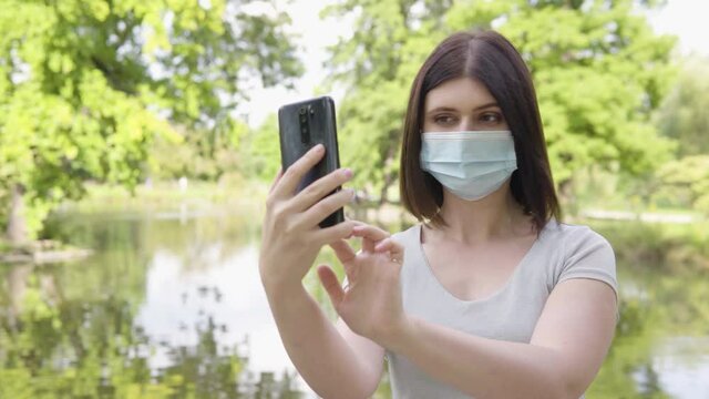 A young Caucasian woman in a face mask takes selfies with a smartphone in a park - a pond and trees in the blurry background