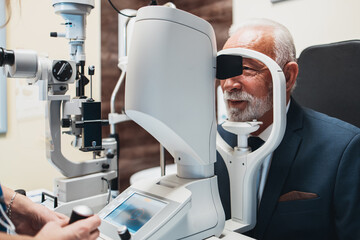 Elegant senior bearded man receiving ophthalmology treatment. Doctor ophthalmologist checking his eyesight with modern equipment. - obrazy, fototapety, plakaty