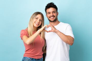 Young caucasian couple isolated on blue background making a heart with hands