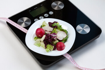 some salad and a pink centimeter on white plate standing on black scales on white background