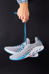 Hand holds hanging gray running sneakers by the laces on black background. Hand with a new sport...