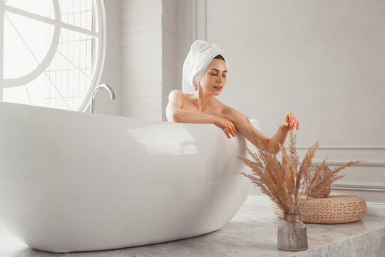 Brunette pretty woman with under-eye patches and towel on head enjoying bathing in spa. Luxiry bathroom interior