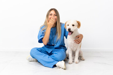 Young veterinarian woman with dog sitting on the floor happy and smiling covering mouth with hand