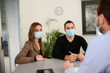 Young couple signing documents in a business bank insurance office wearing  protective surgical mask with a professional agent during covid 19 pandemic