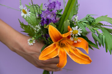 A bouquet of colorful flowers and an orange lily in a woman's hand on a lilac background. The concept of gifts