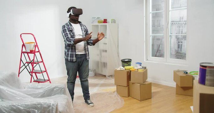 African American young guy stands in room during renovation process in VR glasses using futuristic technology for creating home design, house renovation, designing apartment, virtual reality concept