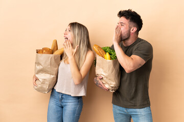 Couple holding grocery shopping bags over isolated background shouting with mouth wide open to the lateral