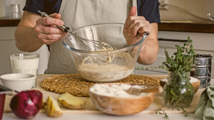 A man without a face in an apron holds a whisk in his hands and kneads dough for baking in the kitchen. Authentic home cooking hobby, home baker. Man cooking in the kitchen at home
