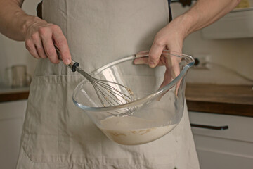 A man without a face in an apron holds a whisk in his hands and kneads dough for baking in the...