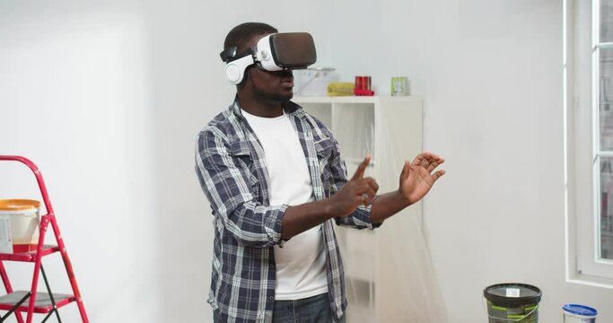 Portrait of African American young guy stands in room in VR glasses using futuristic technology moving hands in air creating home design, house renovation, designing apartment, virtual reality concept