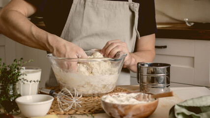 A faceless man in an apron in his kitchen at the kitchen table kneads bread dough in a glass bowl with his hands. Home authentic hobby, home baker. Baking bread with your own hands